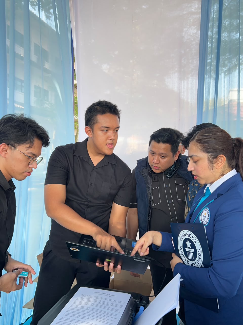 Empat orang, tiga pria dan seorang wanita, berdiri mengelilingi sebuah meja dan laptop yang terbuka di lingkungan semi-outdoor. Seorang pria berbaju hitam menunjuk ke layar laptop. Wanita di sebelah kanan, mengenakan blazer biru dengan logo 'GUINNESS
