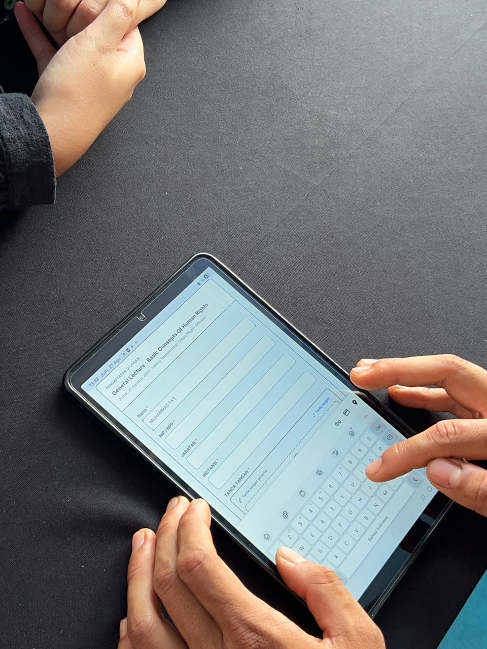 Tampilan jarak dekat tangan seseorang yang sedang mengetik pada keyboard virtual di layar tablet. Layar tablet menampilkan formulir pendaftaran digital berbahasa Indonesia untuk acara "General Lecture: Basic Concepts of Human Rights". Tablet diletakk