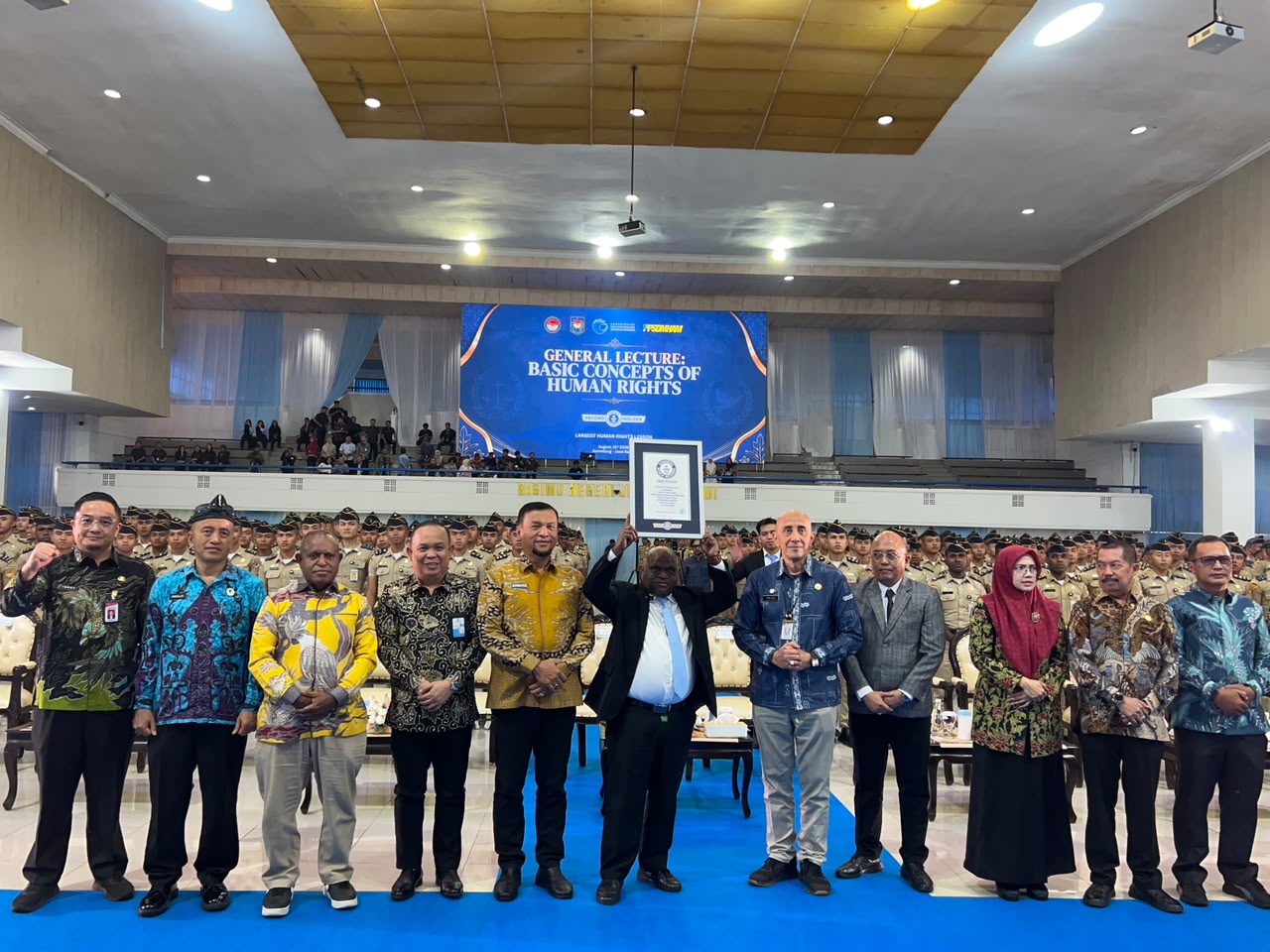 Pemandangan di panggung utama sebuah aula besar yang dihadiri ratusan penonton berseragam di latar belakang. Sekelompok pejabat tinggi berpose untuk foto bersama. Pria di tengah memegang sertifikat rekor besar. Layar LED di panggung menampilkan teks