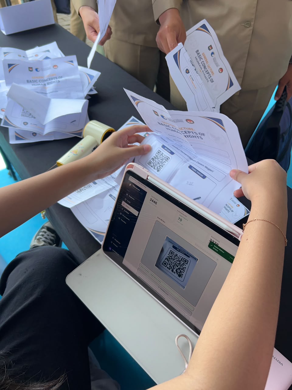 Pandangan dari atas bahu seseorang yang sedang menggunakan tablet bersampul putih untuk memindai QR code pada lembar tiket cetak. Layar tablet menampilkan antarmuka kamera pemindai dari sistem PintuAcara. Di depannya, terlihat tangan peserta berserag