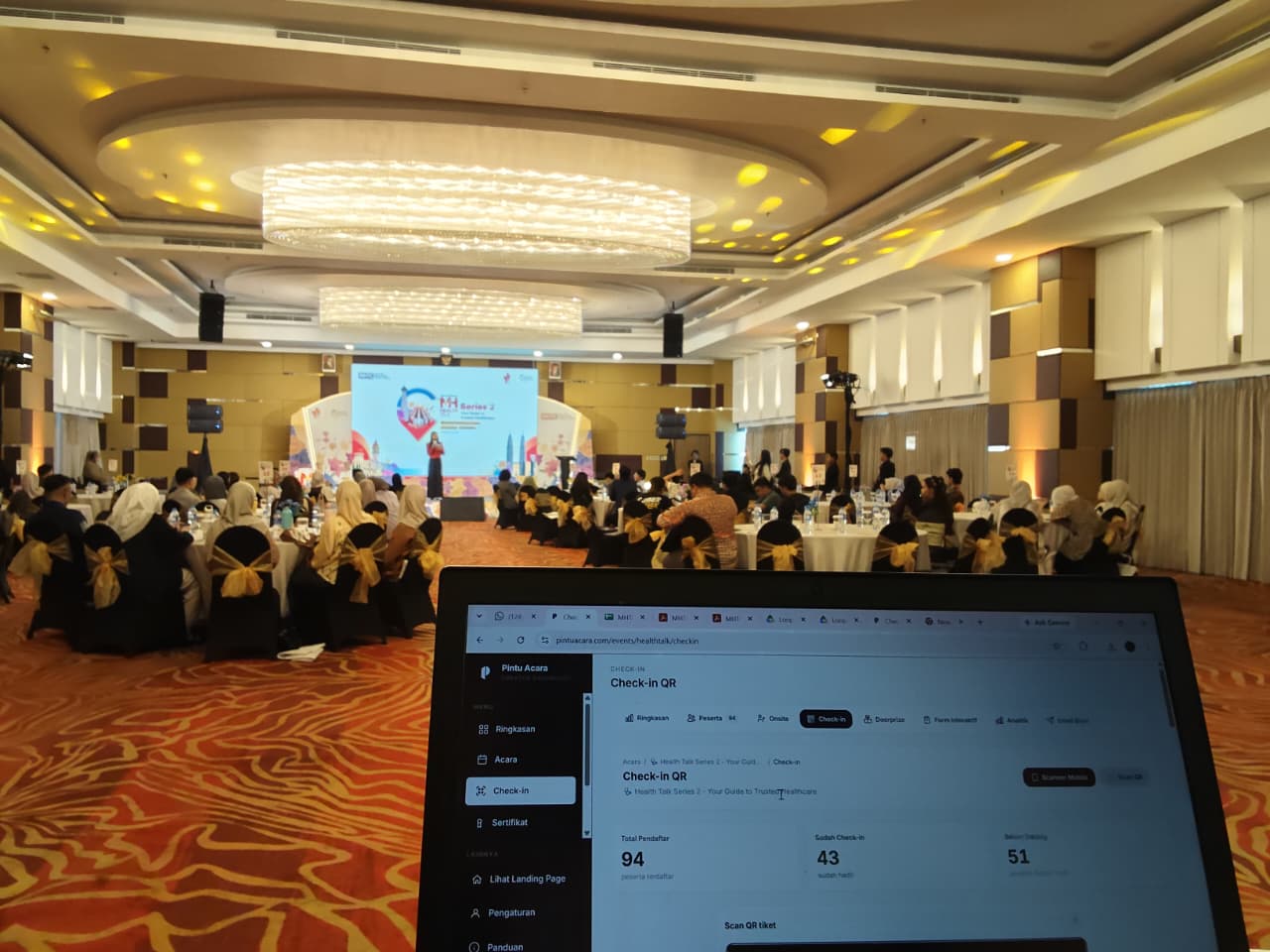 Latar depan laptop menampilkan dasbor check-in Pintu Acara di acara MHTC Health Talk Makassar, dengan latar belakang penonton di ballroom.
